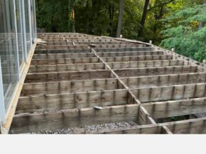 Wooden deck framing under construction next to a glass-walled structure in a wooded area of Rochester, NY.