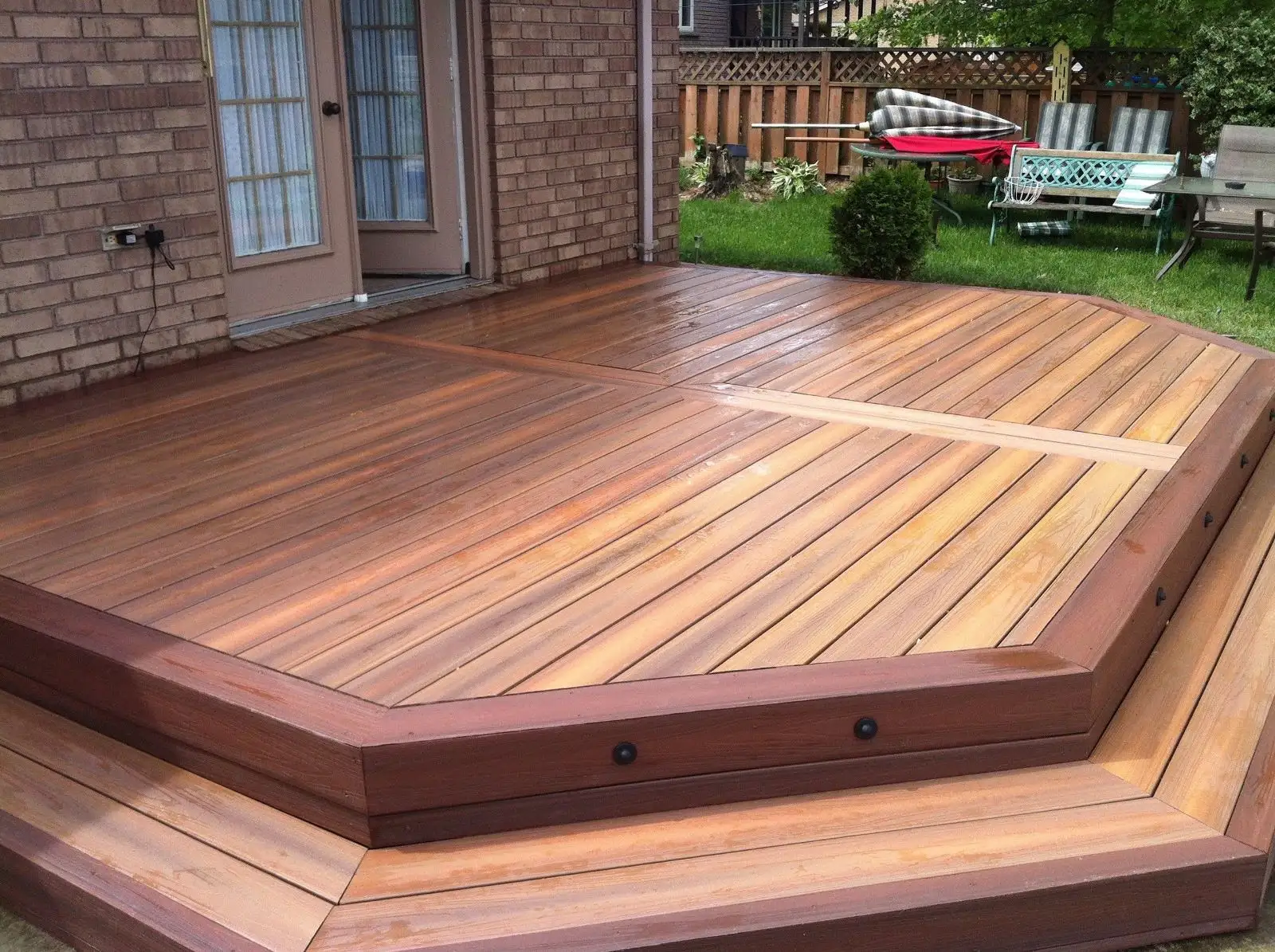 A wet, multi-level wooden deck with a curved edge is attached to a brick house. Steps with dark trim lead down to the main deck area.