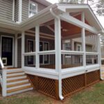 A newly constructed screened-in porch with white trim, black railings, and brown lattice skirting is attached to a light brown house. Wooden stairs with white railings lead up to the porch and down to the yard.