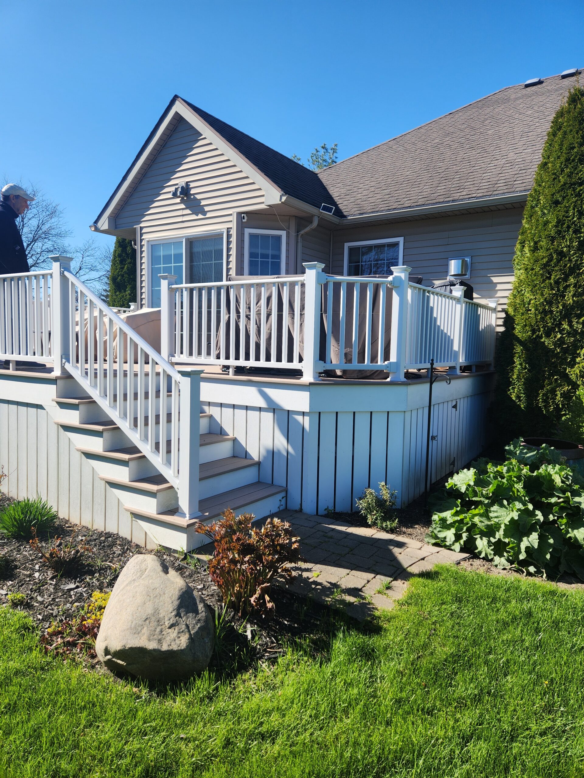 A beautiful elevated wood deck or composite deck in Rochester NY with white vinyl railings, wide stairs, and vertical skirting, attached to a home with light siding, built by a skilled deck builder.
