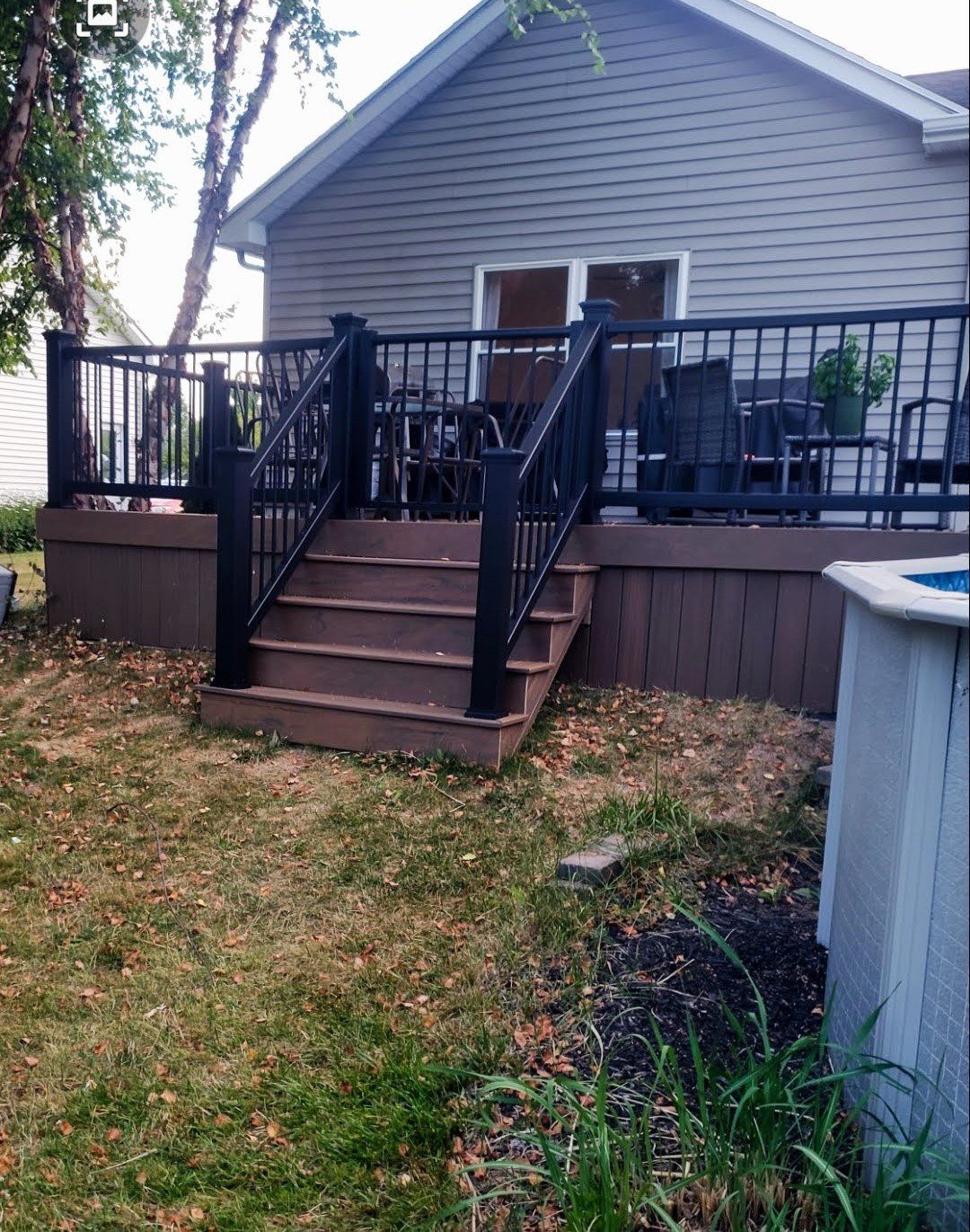 An elevated dark brown composite deck in Rochester NY featuring black aluminum railings and central deck stairs leading to the yard, with a partial pool view, installed by a professional deck builder.