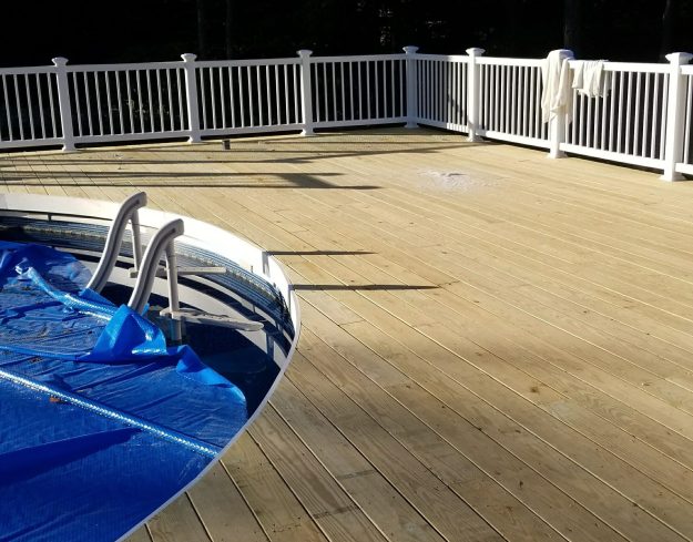 A spacious light-colored wood deck in Rochester NY with white vinyl railings surrounding an above-ground pool, built by a professional deck builder.