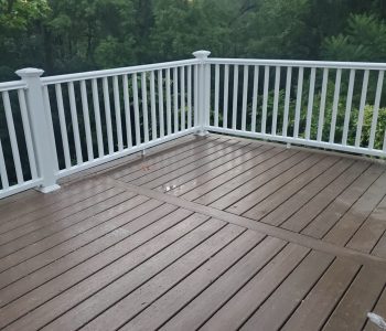 A durable brown composite deck in Rochester NY featuring crisp white vinyl railings, shown wet after rain with a lush tree backdrop, expertly installed by a local deck builder.
