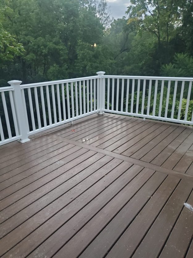 A durable brown composite deck in Rochester NY featuring crisp white vinyl railings, shown wet after rain with a lush tree backdrop, expertly installed by a local deck builder.