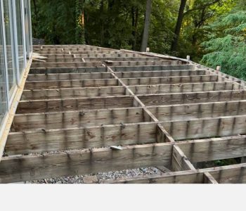 Wooden deck framing under construction next to a glass-walled structure in a wooded area of Rochester, NY.