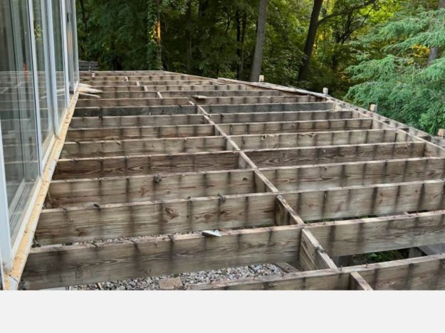 Wooden deck framing under construction next to a glass-walled structure in a wooded area of Rochester, NY.