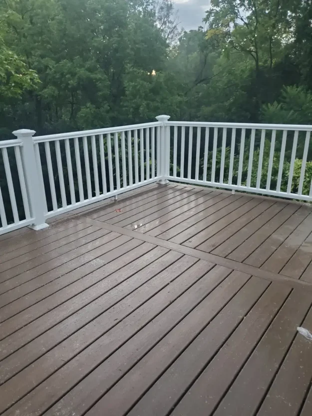 Brown composite deck with white vinyl railings in a wooded Rochester NY backyard, shown wet after rain, built by local decking contractors.